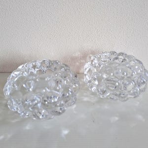 May include: Two clear glass candle holders with a bubbly, textured design. The candle holders are round and sit on a white surface. The background is a textured, off-white wall.