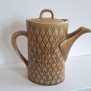 Peut inclure: Une théière cylindrique en céramique avec un vernis brun moutarde, ornée d'un motif de feuilles en relief sur tout le corps. Elle comprend un couvercle assorti avec une petite anse arquée, une poignée latérale courbée et un bec verseur large.
