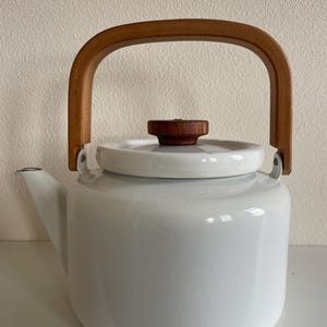Puede incluir: Tetera de esmalte blanco con asa y pomo de madera. La tetera tiene un cuerpo redondeado, un pico y un asa curva de madera. La tapa es blanca con un pequeño pomo redondo de madera.