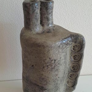 May include: A tall, hand-built ceramic vase with a mottled, earthy gray-brown finish. The vessel features two distinct cylindrical necks at the top and a vertical side panel decorated with a row of five circular, spiral-patterned indentations.