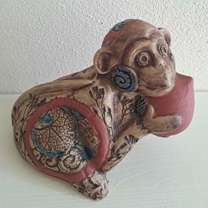 Puede incluir: Una figura de mono de cerámica en tonos beige, marrón y rojo terracota. La superficie presenta intrincados patrones florales grabados, un recorte circular con textura de hoja y una esfera roja lisa sostenida por el mono con sus patas delanteras.