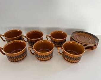 May include: Set of six brown and black teacups with a geometric pattern. The cups are arranged in a semi-circle on a white surface. There is a matching saucer in the background.