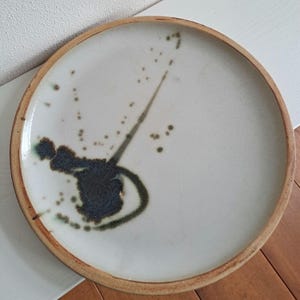 Puede incluir: Un plato redondo de cerámica con una superficie de color crema y un borde marrón. El plato presenta un diseño abstracto azul oscuro y verde con salpicaduras y una línea curva. El plato es adecuado para servir alimentos.