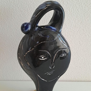 Könnte beinhalten: Eine glänzend schwarze Keramikvase in Form eines stilisierten menschlichen Kopfes mit einem großen, bogenförmigen Henkel an der Oberseite. Das Gesicht zeigt eingravierte Linien für Augen, Nase und Mund sowie eine kleine gelb-grüne Blume am Hals.