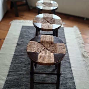 Puede incluir: Tres taburetes redondos de madera dispuestos en línea, cada uno con una estructura de madera marrón oscuro y un asiento circular con un centro de fibras naturales tejidas. Los taburetes tienen cuatro patas unidas por travesaños.