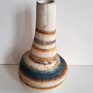 Può includere: Un vaso in ceramica con collo cilindrico e base bulbosa. Il vaso presenta fasce orizzontali di colore, tra cui marrone, verde acqua e crema. Il vaso è alto circa 25 cm.