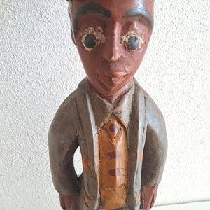 May include: A hand-carved wooden figurine of a person wearing a light-coloured pith helmet, a grey jacket, an orange waistcoat with brown accents, and dark trousers. The figure stands on a circular base and features painted facial details and eyes.