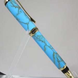 Puede incluir: Un bol&iacute;grafo turquesa y dorado con un dise&ntilde;o veteado. El bol&iacute;grafo tiene detalles dorados en el clip, la punta y el capuch&oacute;n. Se muestra sobre un soporte de acr&iacute;lico transparente.