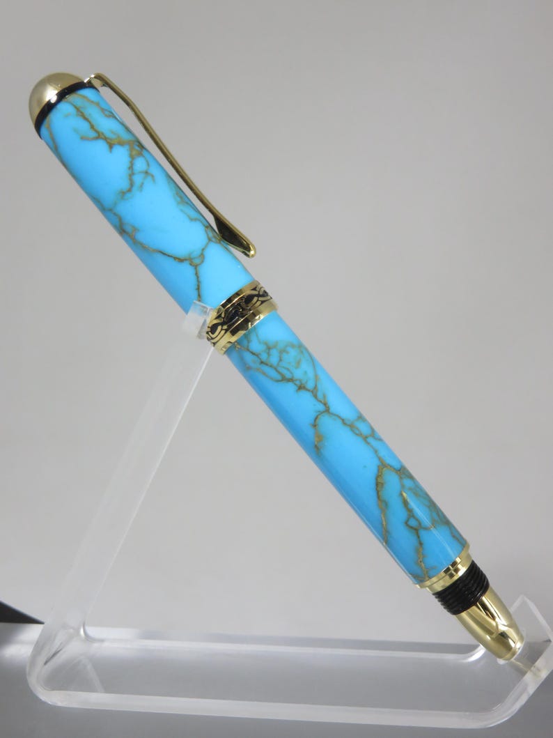 Puede incluir: Un bol&iacute;grafo turquesa y dorado con un dise&ntilde;o veteado. El bol&iacute;grafo tiene detalles dorados en el clip, la punta y el capuch&oacute;n. Se muestra sobre un soporte de acr&iacute;lico transparente.