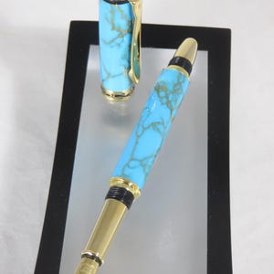 Puede incluir: Una pluma estilogr&aacute;fica con un cuerpo turquesa veteado y detalles dorados. La tapa de la pluma est&aacute; separada y colocada junto a la pluma. La pluma se muestra sobre una superficie reflectante dentro de un marco negro.