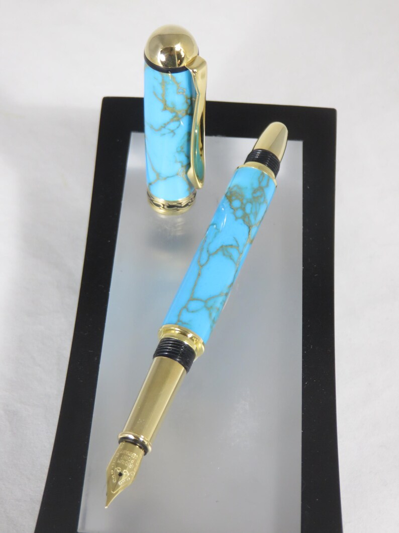 Puede incluir: Una pluma estilogr&aacute;fica con un cuerpo turquesa veteado y detalles dorados. La tapa de la pluma est&aacute; separada y colocada junto a la pluma. La pluma se muestra sobre una superficie reflectante dentro de un marco negro.