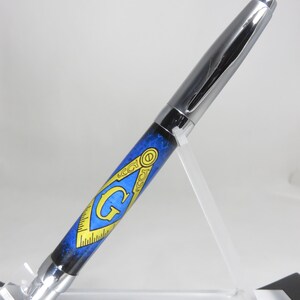 May include: A silver pen with a blue and black design featuring a gold Masonic symbol with a 'G' in the center. The pen is displayed on a clear acrylic stand.