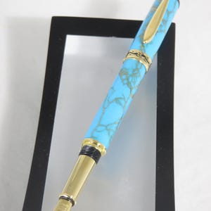 Puede incluir: Una pluma estilogr&aacute;fica turquesa y dorada. El bol&iacute;grafo presenta un cuerpo azul claro con un patr&oacute;n veteado, detalles dorados y un plum&iacute;n dorado. El bol&iacute;grafo se muestra sobre un fondo blanco.