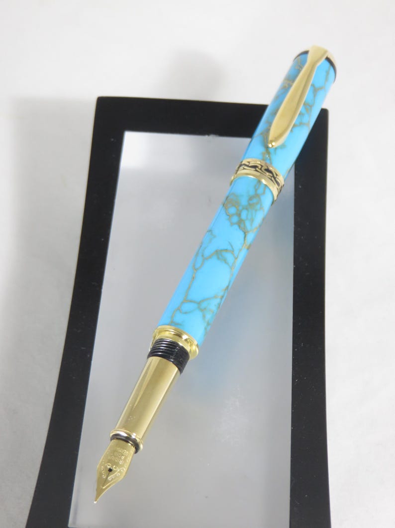 Puede incluir: Una pluma estilogr&aacute;fica turquesa y dorada. El bol&iacute;grafo presenta un cuerpo azul claro con un patr&oacute;n veteado, detalles dorados y un plum&iacute;n dorado. El bol&iacute;grafo se muestra sobre un fondo blanco.