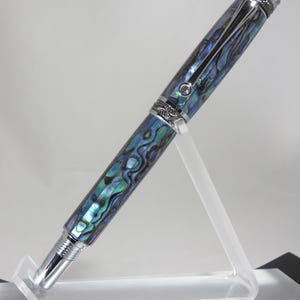 May include: A luxury fountain pen with a shimmering abalone shell design in shades of blue and green. The pen has silver-coloured metal accents and a decorative cap. The pen is displayed on a clear acrylic stand.