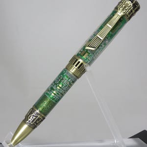 May include: A unique pen with a circuit board design. The pen features a brass-colored tip and accents, with a green and gold circuit board pattern on the barrel. The pen is displayed on a clear acrylic stand.