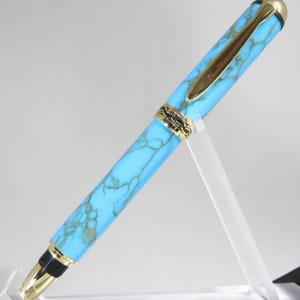 Puede incluir: Una pluma estilogr&aacute;fica turquesa y dorada. La pluma presenta un cuerpo turquesa veteado con detalles dorados, incluyendo el clip, el plum&iacute;n y las bandas decorativas. La pluma se exhibe sobre un soporte de acr&iacute;lico transparente.