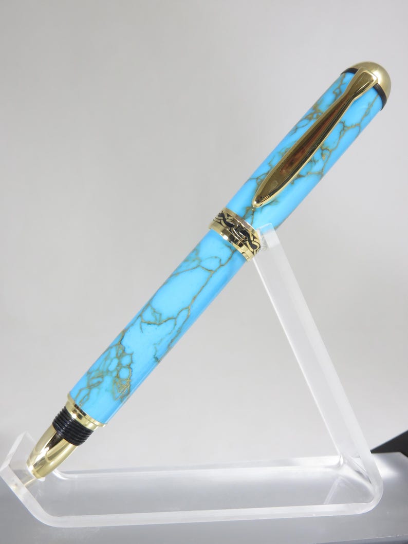 Puede incluir: Una pluma estilogr&aacute;fica turquesa y dorada. La pluma presenta un cuerpo turquesa veteado con detalles dorados, incluyendo el clip, el plum&iacute;n y las bandas decorativas. La pluma se exhibe sobre un soporte de acr&iacute;lico transparente.