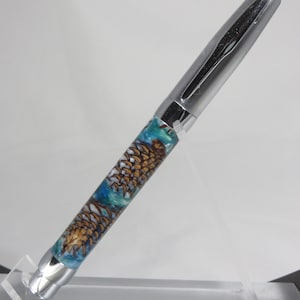 Peut inclure: Un stylo argenté avec un corps en résine bleu et brun orné d'un motif de pomme de pin. Le stylo est présenté sur un support en acrylique transparent.