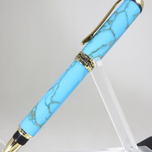 Puede incluir: Un bol&iacute;grafo turquesa y dorado con un dise&ntilde;o veteado. El bol&iacute;grafo tiene un clip y detalles dorados. El bol&iacute;grafo se exhibe en un soporte de acr&iacute;lico transparente. Es un instrumento de escritura.