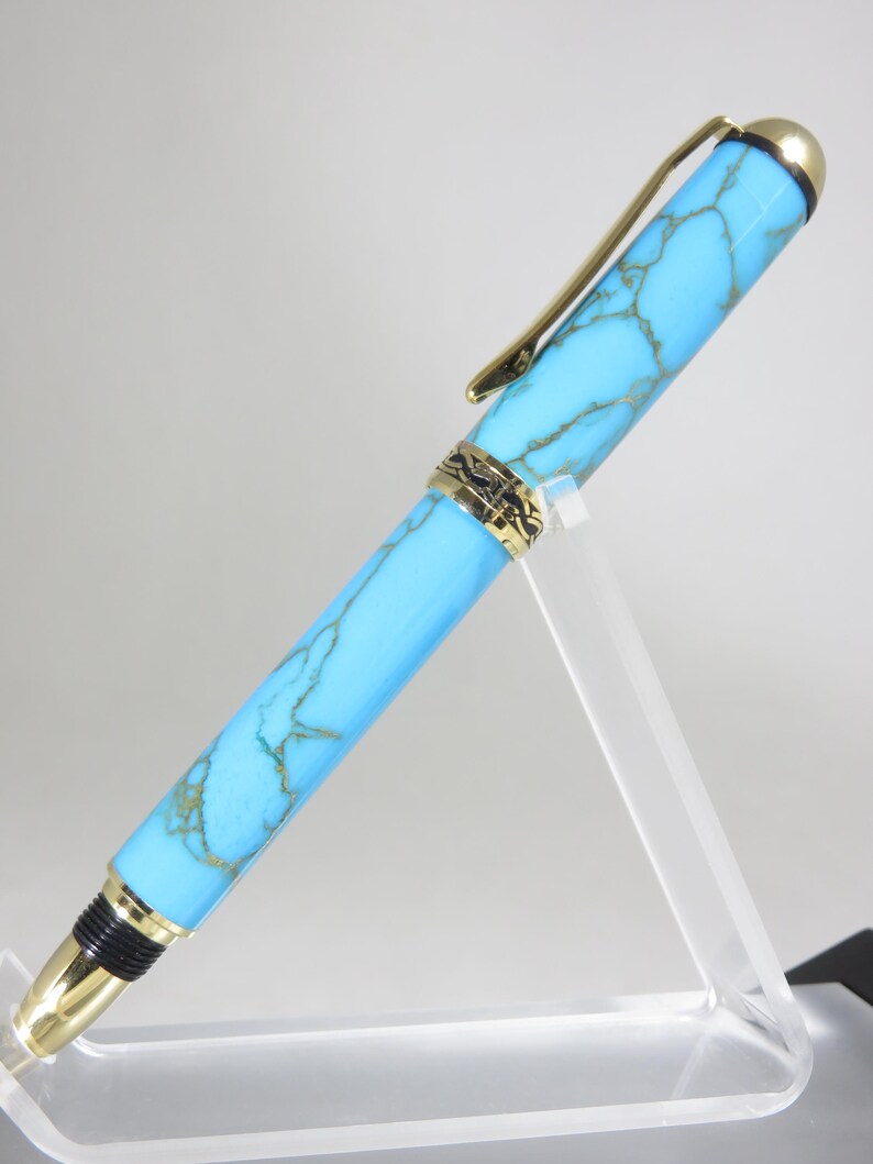 Puede incluir: Un bol&iacute;grafo turquesa y dorado con un dise&ntilde;o veteado. El bol&iacute;grafo tiene un clip y detalles dorados. El bol&iacute;grafo se exhibe en un soporte de acr&iacute;lico transparente. Es un instrumento de escritura.