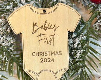 Op de afbeelding: Een houten kerstornament in de vorm van een babyromper. Het lichtbruine ornament heeft de gegraveerde tekst "Babies First CHRISTMAS 2024". Het is omgeven door groenblijvende takken, rode bessen en een dennenappel.
