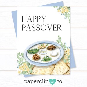 Puede incluir: Una tarjeta de felicitación con las palabras "HAPPY PASSOVER" en texto negro. La tarjeta presenta una ilustración en acuarela de un plato de Seder con alimentos tradicionales, rodeada de detalles florales. La tarjeta está sobre un fondo azul claro.