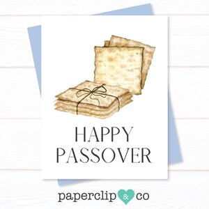 May include: A white greeting card with a light blue envelope. The card features a watercolor illustration of matzah crackers tied with twine. The words "HAPPY PASSOVER" are printed in black. The card also has the text "paperclip & co".