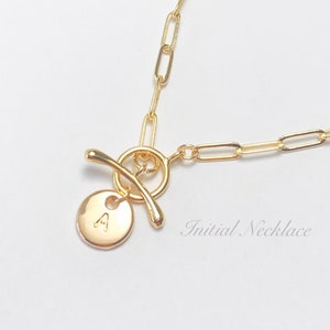 Puede incluir: Un collar de cadena dorada con un cierre de palanca y un colgante redondo con la letra "A" grabada. El collar está sobre un fondo blanco. "Initial Necklace" está escrito en la esquina inferior derecha de la imagen.