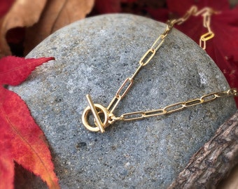 May include: A gold chain necklace with a toggle clasp. The chain is made of small, rectangular links. The necklace is lying on a grey rock.