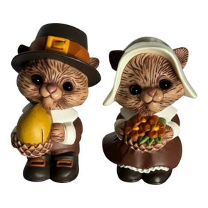 May include: Two ceramic cat figurines dressed in historical attire. The left cat wears a brown hat with a yellow buckle and holds a yellow gourd. The right cat wears a white bonnet and holds a basket of orange flowers. Both have brown fur.