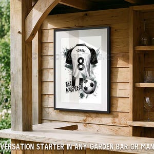 May include: Framed print of a black and white soccer jersey with the name "TONALI" and the number "8". A soccer ball and the words "THE MAGPIES" are also featured. The print is displayed on a wooden wall.