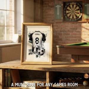 May include: Framed artwork featuring a soccer jersey with the number 8 and the name TONALI. The jersey is black and white, with a soccer ball and the text "THE MAGPIES". The artwork is displayed on a wooden table.
