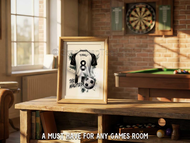 May include: Framed artwork featuring a soccer jersey with the number 8 and the name TONALI. The jersey is black and white, with a soccer ball and the text "THE MAGPIES". The artwork is displayed on a wooden table.