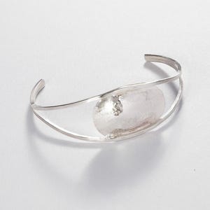 May include: A silver cuff bracelet featuring a hammered, oval centrepiece. The bracelet has a minimalist design with a polished finish, suitable for everyday wear. The open design allows for easy adjustment and comfortable fit.