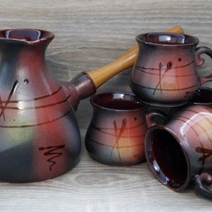 May include: A ceramic coffee set with a dark brown, red, and gray color scheme. The set includes a coffee pot with a wooden handle and four matching mugs. The pieces have a hand-painted design with abstract lines.