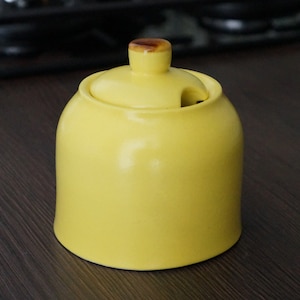 May include: A yellow ceramic sugar bowl with a lid and a small handle. The lid has a slot for a spoon. The handle is brown. The bowl is sitting on a dark wooden surface.
