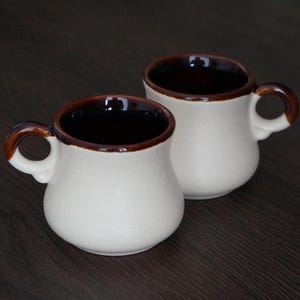 May include: Two ceramic mugs with a cream-colored exterior and a dark brown interior and handle. The mugs have a rounded shape and are set against a dark wood background. The mugs are designed for hot beverages.