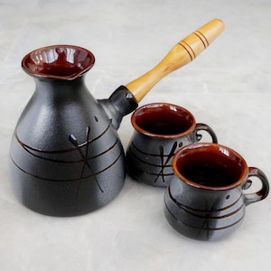 May include: A ceramic coffee pot with a long wooden handle and two matching small cups. The set features a matte black exterior with a glossy dark red interior and simple etched line patterns on the rounded bodies of the pot and the cups.
