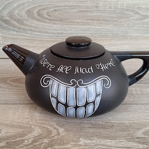 May include: A matte black ceramic teapot featuring a white hand-painted graphic of a wide, toothy grin and the text "We're all mad here" written in a cursive script above the mouth. The teapot includes a matching lid, a curved handle, and a spout.