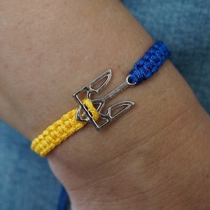 May include: A silver trident charm bracelet with yellow and blue woven bands. The bracelet features a Ukrainian trident symbol, with yellow and blue representing the Ukrainian flag. The bracelet is worn on a wrist.