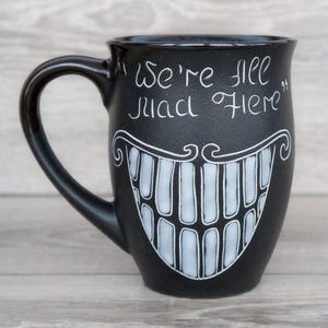 May include: A black ceramic mug featuring a white hand-drawn illustration of a wide, toothy grin and the text "We're all mad here" written in a stylized script font above the mouth. The mug has a curved handle and a smooth, matte finish.