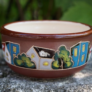 May include: A brown ceramic bowl with a cream-colored interior. The exterior features a painted village scene with houses, trees, and blue and yellow accents. The bowl is sitting on a stone surface.