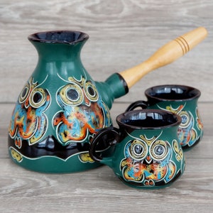 May include: A teal ceramic coffee pot with a wooden handle and two matching mugs. Each piece is decorated with a colorful owl design. The set is designed for brewing and serving coffee.
