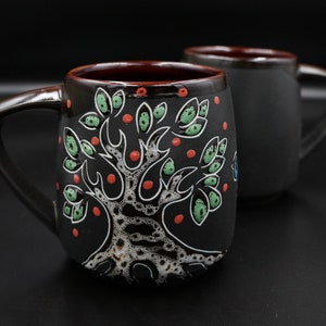 May include: Two black ceramic mugs with handles. One mug has a white tree design with green leaves and red dots. The other mug is plain.