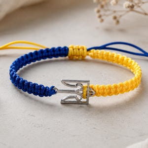 May include: A braided bracelet in blue and yellow, featuring a silver trident charm. The adjustable bracelet is made of woven cord, with the colors representing the Ukrainian flag. The bracelet is a symbol of solidarity and support.