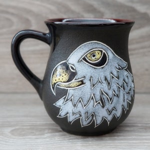May include: A black ceramic mug with a handle, featuring a detailed illustration of an eagle's head. The eagle's head is white and gray with yellow accents, set against the dark background of the mug. The mug has a textured surface.