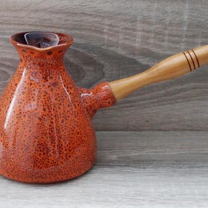 May include: A ceramic coffee pot with a wide base and a narrow neck, featuring a speckled orange and brown glaze. The pot has a long, light-colored wooden handle attached to the side, which is decorated with three thin, dark horizontal lines.