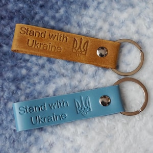 May include: Two leather keychains with silver key rings. Both keychains are blue and brown and feature the text "Stand with Ukraine" and the Ukrainian trident symbol.