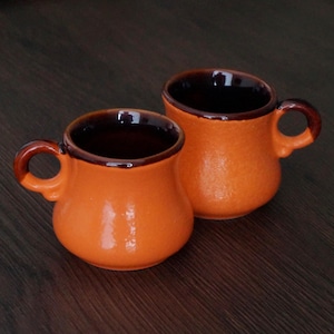 May include: Two small, rounded ceramic mugs with a bright orange exterior and a dark brown glazed interior and rim. Each mug features a curved handle attached to the side, with a smooth, glossy finish on the rim and a textured orange body.